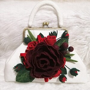 Puede incluir: Una cartera de mano blanca de fieltro con un cierre dorado y una gran rosa de fieltro burdeos con rosas de fieltro rojo y hojas de fieltro verde.