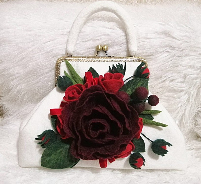 Puede incluir: Una cartera de mano blanca de fieltro con un cierre dorado y una gran rosa de fieltro burdeos con rosas de fieltro rojo y hojas de fieltro verde.