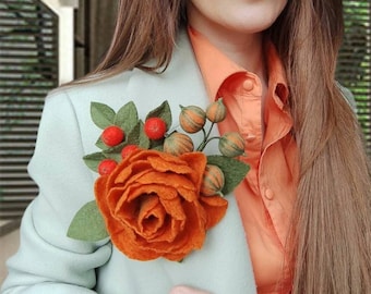 以下が含まれることがあります： オレンジの花びらと緑の葉を持つフェルトの花のブローチ。ブローチには小さなオレンジ色のベリーと緑のさやが付いています。ブローチは薄いブルーのブレザーにピンで留められています。