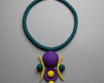 May include: A felted necklace featuring a thick teal circular band with a silver-toned clasp. The pendant consists of a large purple sphere, a smaller purple disc with a yellow center, two teal side spheres, two yellow crescent shapes, and a yellow sphere at the base.