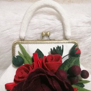 Puede incluir: Un bolso de mano de fieltro blanco con cierre dorado y asa de fieltro blanco. El bolso est&aacute; decorado con un grupo de rosas de fieltro rojo y burdeos y hojas de fieltro verde.