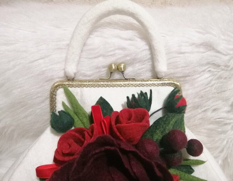 Puede incluir: Un bolso de mano de fieltro blanco con cierre dorado y asa de fieltro blanco. El bolso est&aacute; decorado con un grupo de rosas de fieltro rojo y burdeos y hojas de fieltro verde.