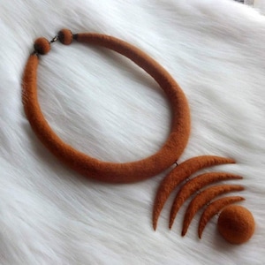 May include: A brown felt necklace with a large, round pendant made of multiple crescent shapes. The pendant is attached to the necklace with a small metal ring.