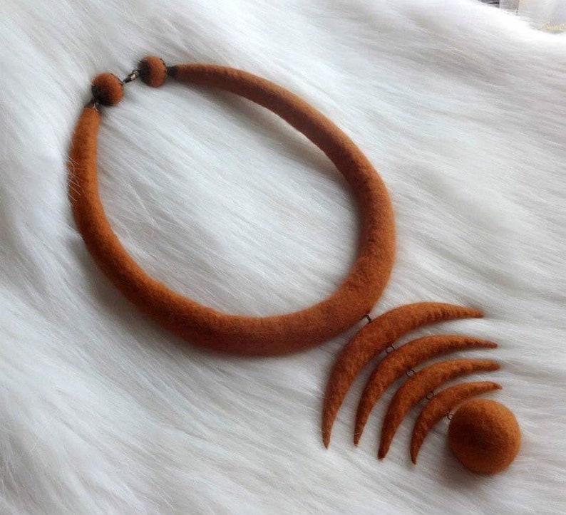May include: A brown felt necklace with a large, round pendant made of multiple crescent shapes. The pendant is attached to the necklace with a small metal ring.