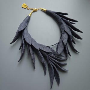 Puede incluir: Un collar de fieltro gris con un diseño en forma de hojas. El collar tiene una cadena y un cierre dorados. Las hojas están dispuestas en un patrón en capas, creando un aspecto texturizado. El collar se muestra sobre un fondo gris.