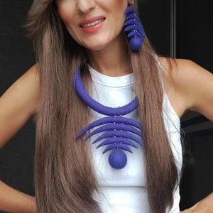 May include: A woman wearing a white tank top with a large, geometric, purple felt necklace and matching earrings.
