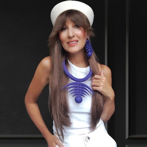 May include: A woman wearing a white hat, white top, and white pants. She is wearing a large, purple felt necklace and matching earrings.