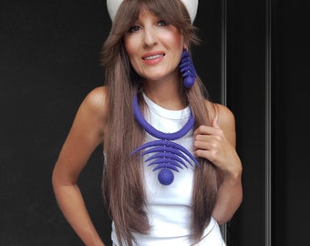 May include: A woman wearing a white hat, white top, and white pants. She is wearing a large, purple felt necklace and matching earrings.