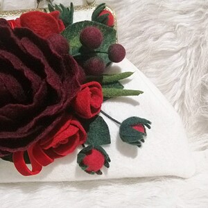 Puede incluir: Un bolso de mano blanco de fieltro con un arreglo floral de fieltro burdeos y rojo. Las flores est&aacute;n hechas de fieltro y tienen un aspecto realista. El bolso tiene un cierre de metal dorado.