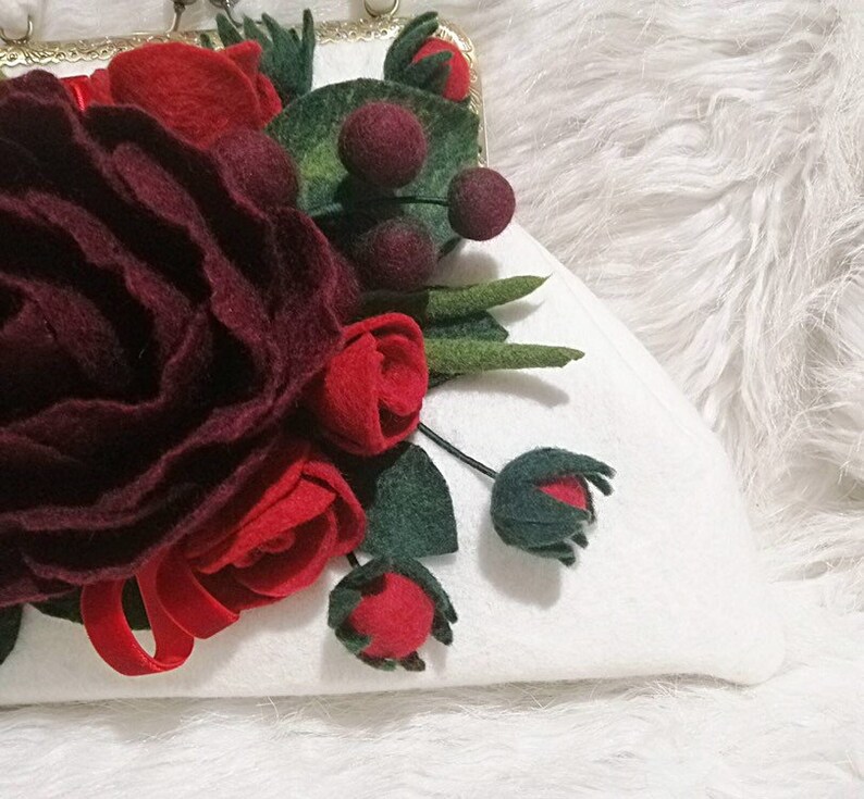 Puede incluir: Un bolso de mano blanco de fieltro con un arreglo floral de fieltro burdeos y rojo. Las flores est&aacute;n hechas de fieltro y tienen un aspecto realista. El bolso tiene un cierre de metal dorado.