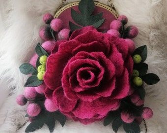 以下が含まれることがあります： フェルト製の丸い小さなクラッチバッグ。フェルトの花と緑の葉でできたバラが飾られています。バッグは金色の金属フレームとチェーンストラップが付いています。