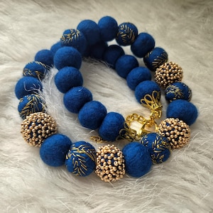 Op de afbeelding: Een handgemaakte ketting met diepblauwe vilten wollen kralen, afgewisseld met goudkleurige kralenaccenten en details van bladgoud. De ketting is vastgezet met een goudkleurige sluiting.