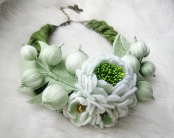 Può includere: Una collana di fiori in feltro bianco con foglie in feltro verde e bacche in feltro bianco. La collana è realizzata con un cordoncino in feltro verde.