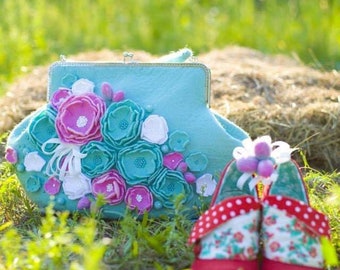 May include: A light blue felt clutch purse with a floral design made of felt flowers in pink, white, and teal. The purse is sitting on a bed of green grass with two red felt shoes with floral and polka dot patterns.