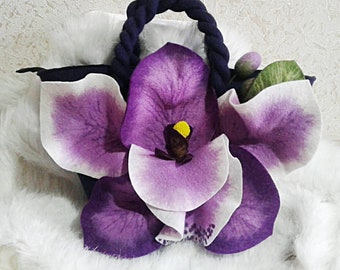 May include: A felted handbag featuring a large, vibrant purple and white orchid. The bag has a navy blue rope handle and is set against a fluffy white background. The orchid has a yellow center and green leaves.