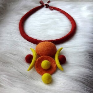 May include: A felted necklace featuring a thick, dark red circular cord with a brown ribbon tie. The pendant consists of a large orange sphere, a flat orange disc, two yellow crescent shapes, two small dark red spheres, and two yellow spheres.