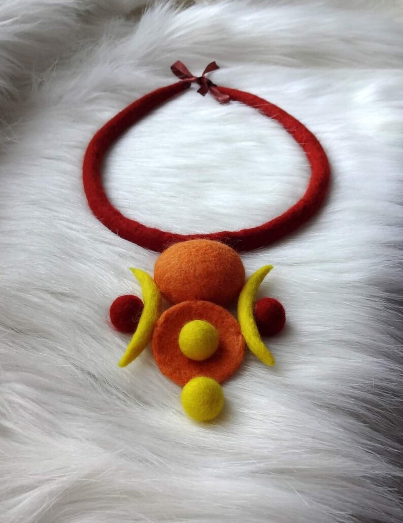 May include: A felted necklace featuring a thick, dark red circular cord with a brown ribbon tie. The pendant consists of a large orange sphere, a flat orange disc, two yellow crescent shapes, two small dark red spheres, and two yellow spheres.