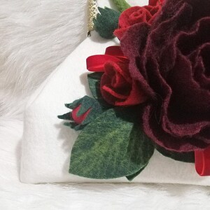 Puede incluir: Una cartera de mano blanca de fieltro con una rosa de fieltro burdeos y hojas de fieltro rojas.