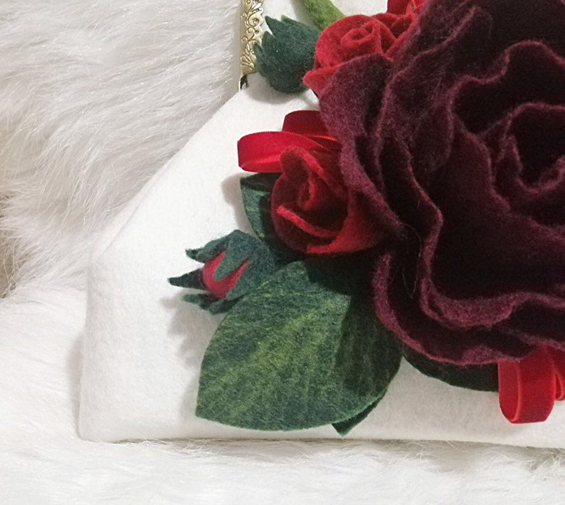 Puede incluir: Una cartera de mano blanca de fieltro con una rosa de fieltro burdeos y hojas de fieltro rojas.