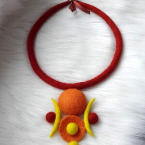 May include: A felt necklace featuring a thick red circular cord tied with a small brown ribbon at the top. The pendant consists of a large orange felt sphere, two yellow crescent shapes, two small red spheres, and two smaller yellow spheres.