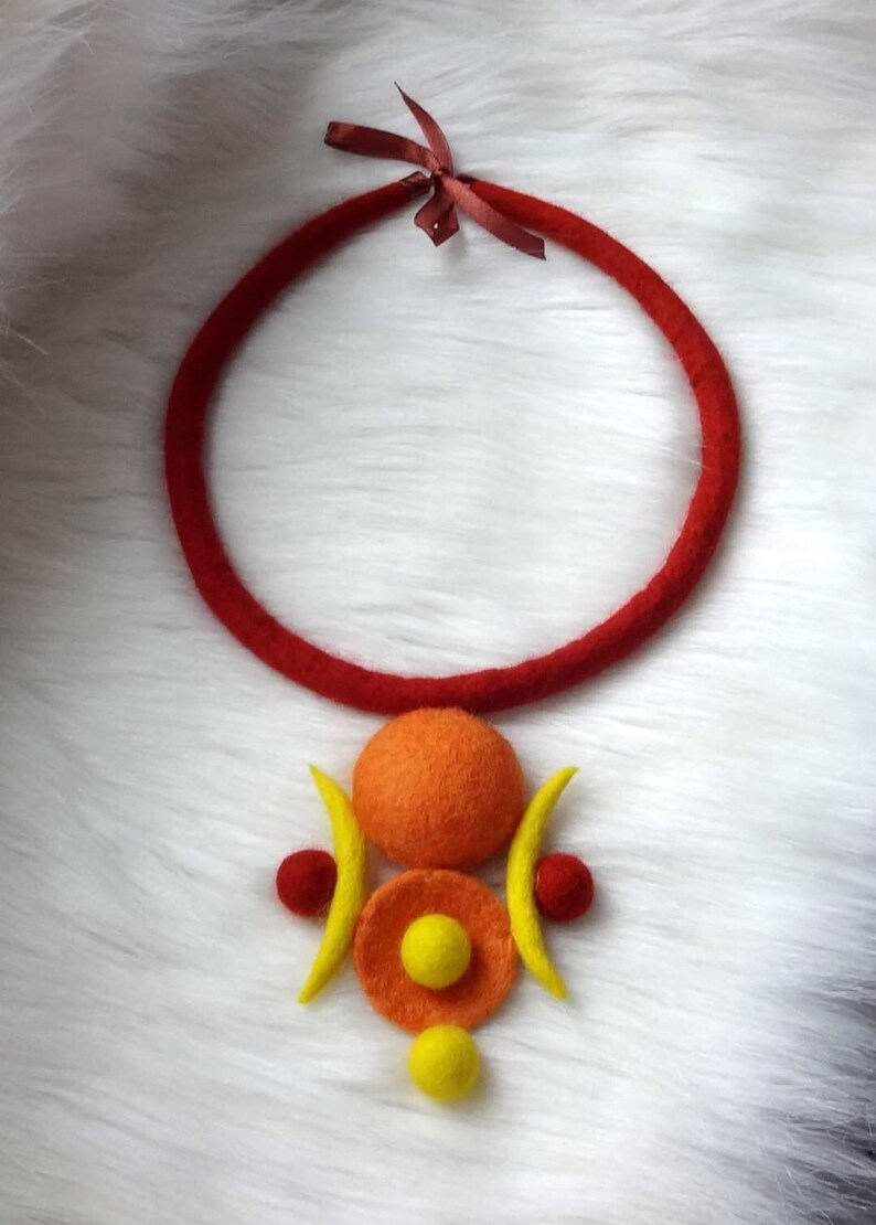 May include: A felt necklace featuring a thick red circular cord tied with a small brown ribbon at the top. The pendant consists of a large orange felt sphere, two yellow crescent shapes, two small red spheres, and two smaller yellow spheres.