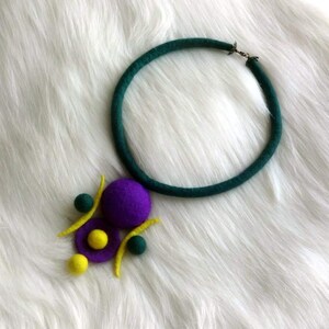 May include: A necklace featuring a dark green felted cord and a pendant composed of purple, yellow, and dark green felted spheres and curved shapes. The pendant is attached to the circular cord, which includes a small metal clasp at the top.