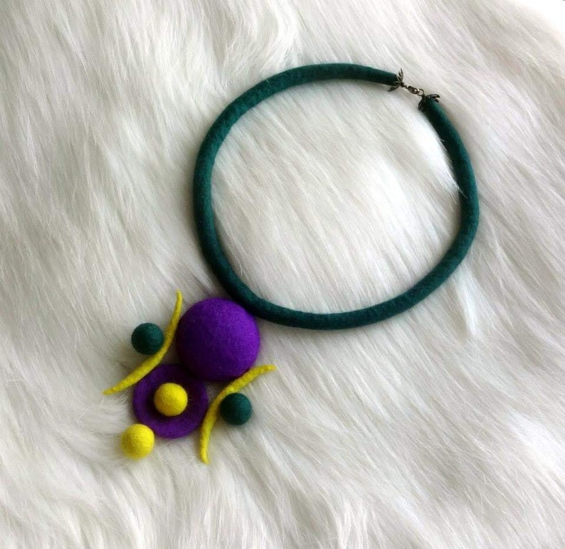May include: A necklace featuring a dark green felted cord and a pendant composed of purple, yellow, and dark green felted spheres and curved shapes. The pendant is attached to the circular cord, which includes a small metal clasp at the top.