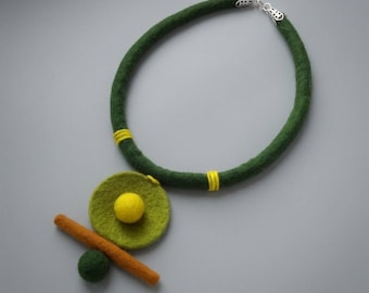 May include: A felt necklace featuring a thick dark green cord with yellow thread accents and a silver-toned clasp. The pendant consists of a lime green felt circle, a bright yellow felt sphere, a dark green felt sphere, and an orange felt rod.
