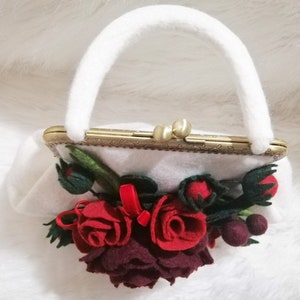Puede incluir: Un bolso de mano blanco de fieltro con un cierre de metal dorado y un asa de fieltro blanco. El bolso est&aacute; decorado con un grupo de flores de fieltro rojo y burdeos.