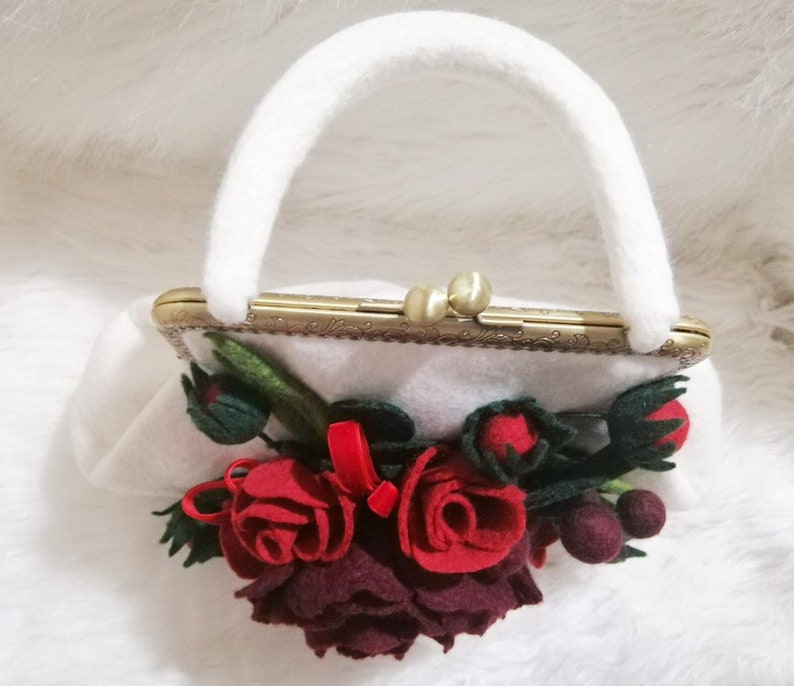 Puede incluir: Un bolso de mano blanco de fieltro con un cierre de metal dorado y un asa de fieltro blanco. El bolso est&aacute; decorado con un grupo de flores de fieltro rojo y burdeos.