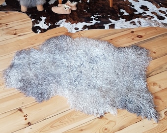 Op de afbeelding: Een lichtgrijs schapenvachtkleed met lange, krullende vacht. Het kleed ligt op een houten vloer met een bruin en wit koeienhuidkleed eronder.