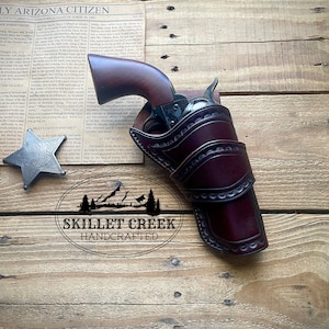 May include: A brown leather holster with a silver-toned gun inside. The holster is decorated with stitching and has a flap that covers the gun. The holster is sitting on a wooden surface. The text "SKILLET CREEK HANDCRAFTED" is visible below the holster.