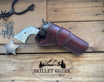 May include: A brown leather holster with intricate stitching and a silver star on a wooden background. The holster is holding a revolver with a white pearl handle. The text "SKILLET CREEK HANDCRAFTED" is visible below the holster.