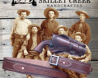 May include: A brown leather gun belt with a silver buckle and a brown leather holster. The holster is designed to hold a revolver. The text "SKILLET CREEK HANDCRAFTED" and "Cheyenne rig" is visible in the image.