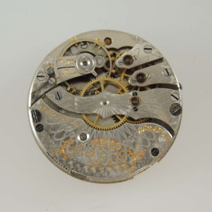 Op de afbeelding: Close-up van een zilveren zakhorloge-uurwerk met ingewikkelde tandwielen en een gouden inscriptie. De wijzerplaat van het horloge ontbreekt.