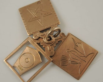 May include: A gold-colored pendant with three hinged sections. One section features a five-pointed star, another has a brush and a quill, and the third has a design within a square frame. The pendant is likely made of metal.
