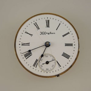 Puede incluir: Una esfera de reloj blanca con marcadores de hora en n&uacute;meros romanos negros y agujas negras. La esfera del reloj tiene la palabra "Hamden" impresa.