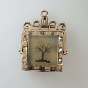 May include: Gold-toned locket pendant with a hinged opening. The locket features a square frame with a decorative border and a clear glass insert. Inside the locket is a silhouette of a tree.