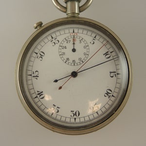 Könnte beinhalten: Eine silberne Vintage-Stoppuhr mit weißem Zifferblatt und schwarzen und roten Zeigern. Die Uhr hat ein kleineres Zifferblatt und einen Ring oben. Die Zahlen sind schwarz, mit einer roten Markierung bei der 3-Sekunden-Marke. Durchmesser ca. 5 cm.