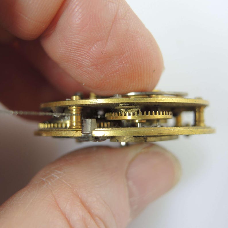 May include: Close-up of a watch mechanism with brass gears and components. The intricate design shows multiple gears meshing together, showcasing the internal workings of a timepiece. The gears are held in place by a metal frame.