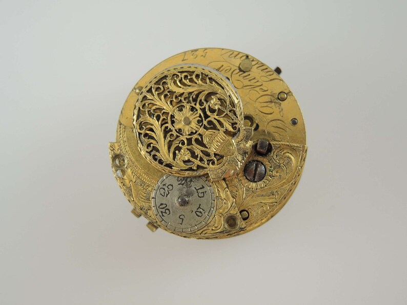 K&ouml;nnte beinhalten: Eine goldfarbene Taschenuhr mit aufwendigen Gravuren und einem filigranen, durchbrochenen Design. Das Zifferblatt der Uhr ist sichtbar und zeigt r&ouml;mische Ziffern und ein kleines, kreisf&ouml;rmiges Zifferblatt.