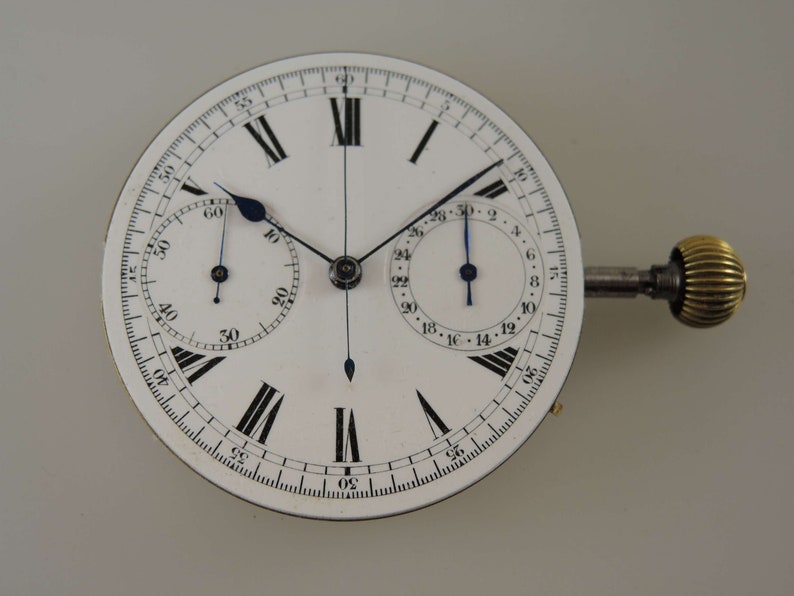 May include: A white chronograph watch face with black Roman numeral hour markers and black minute markers. The watch has two sub-dials, one for seconds and one for minutes. The watch has a gold winding stem.