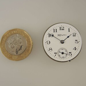 Puede incluir: Una esfera de reloj de bolsillo blanca con marcadores de hora romanos negros y agujas negras. La esfera del reloj dice "The Hampden..." en la parte superior.