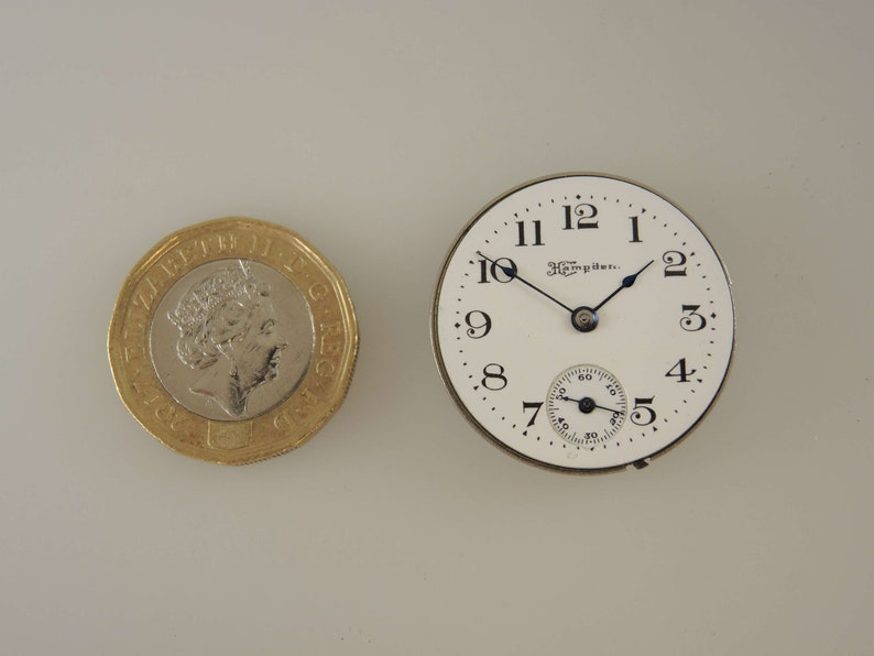 Puede incluir: Una esfera de reloj de bolsillo blanca con marcadores de hora romanos negros y agujas negras. La esfera del reloj dice "The Hampden..." en la parte superior.