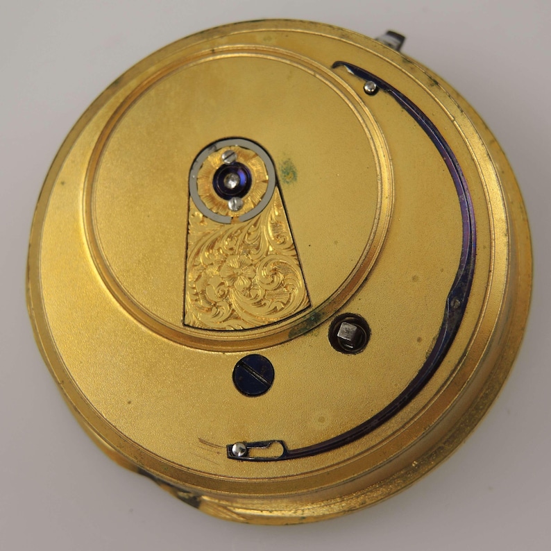 May include: A detailed view of a gold-coloured pocket watch mechanism. The interior showcases intricate engravings and small screws. A curved, dark blue element arcs across the top. The watch's design is circular.