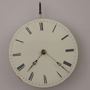 May include: A vintage watch face with a white background and black Roman numerals. The watch face has two delicate hands and a small metal stem at the top. The watch face is round and has a classic design.