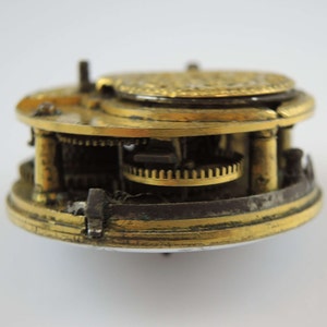 May include: A close-up of a gold-toned pocket watch mechanism with intricate gears and a circular face. The watch is disassembled, revealing the inner workings.