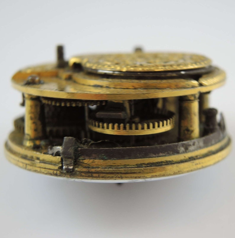 May include: A close-up of a gold-toned pocket watch mechanism with intricate gears and a circular face. The watch is disassembled, revealing the inner workings.
