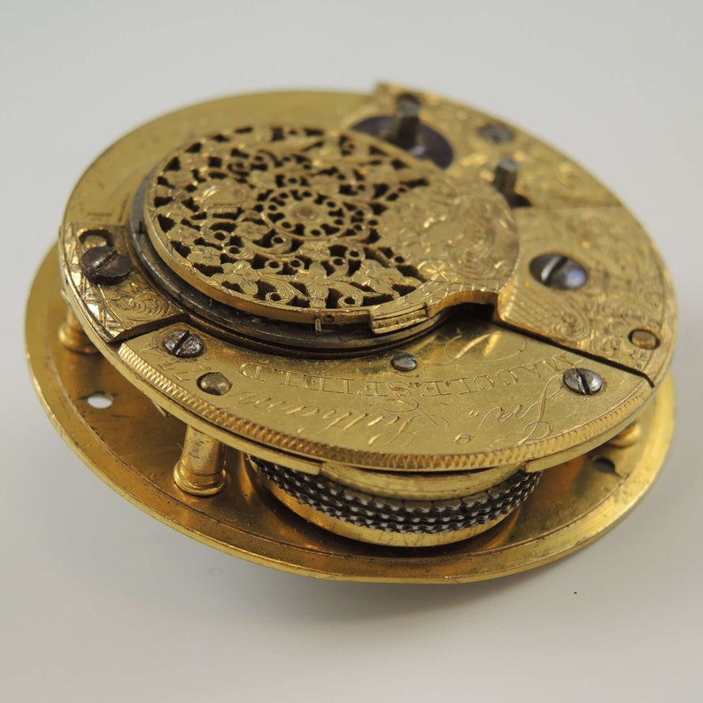 Op de afbeelding: Een gouden zakhorloge met ingewikkelde gravures en een gedetailleerd, opengewerkt ontwerp aan de achterkant. Het horloge heeft een zichtbaar wijzerplaat met de tekst "Thomas Mudge London".