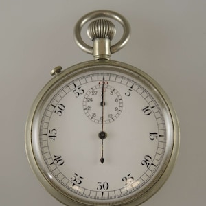 Könnte beinhalten: Eine silberne Taschenuhr mit weißem Zifferblatt und schwarzen Markierungen. Die Uhr hat einen großen Zeiger und einen kleineren Zeiger, der auf die 30-Sekunden-Markierung zeigt. Die Uhr hat eine Kette, die an der Oberseite befestigt ist.